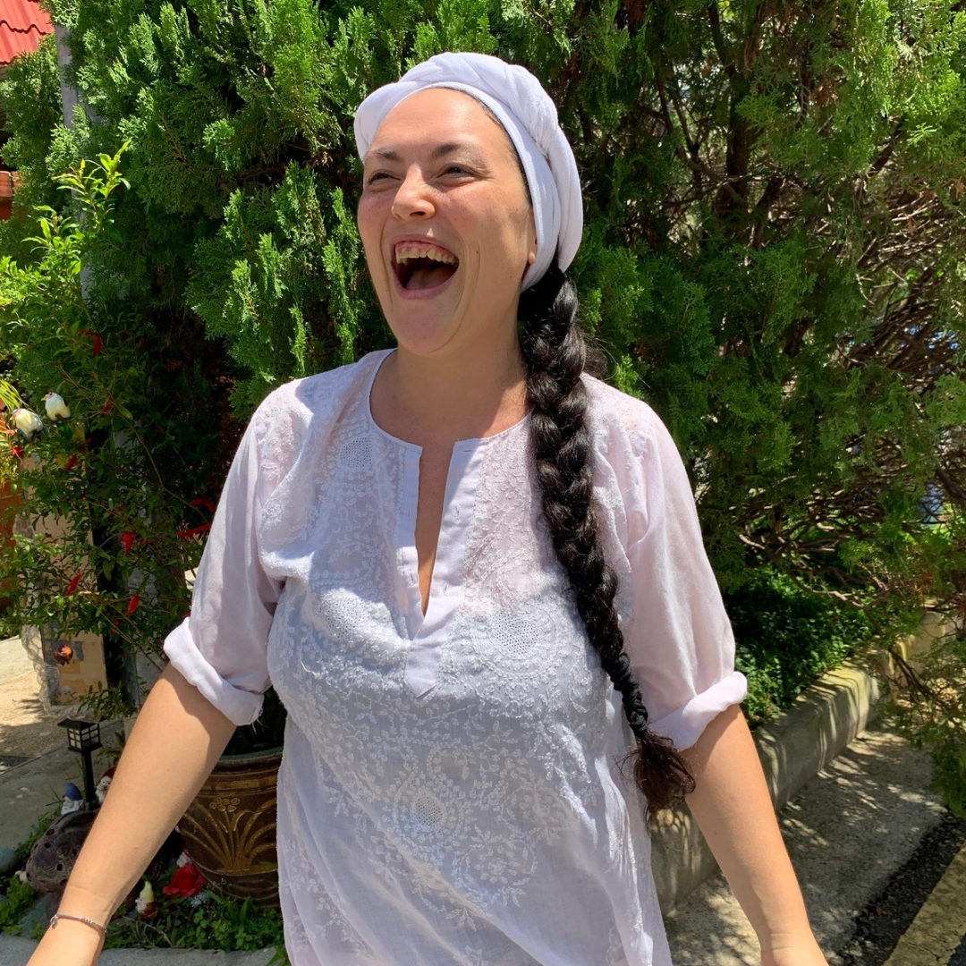 Marilyn, founder of Koru Kundalini, smiling in white yoga clothing against a green garden backdrop